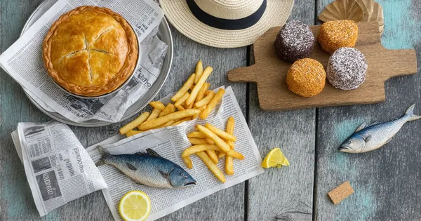 Authentic Australian cuisine with golden meat pie, newspaper-wrapped fish and chips, herb-grilled barramundi, and coconut lamingtons displayed with painted boomerang, Opera House model, and cork hat on weathered beach wood surface